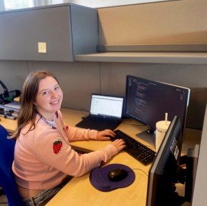 IT student Kenzie smiles at her desk while her computer shows lines of code