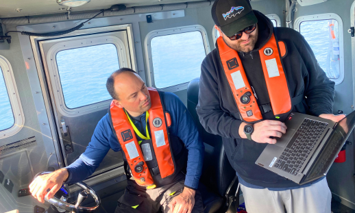 Two men in life jackets aboard the vessel, one shows the other a laptop screen.